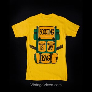 Puede incluir: Camiseta amarilla con una ilustraci&oacute;n verde y naranja de una mochila con el texto "SCOUTING IS MY BAG" en la parte posterior.