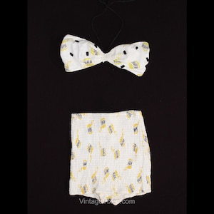 Puede incluir: Traje de baño blanco de dos piezas con estampado geométrico negro y amarillo. La parte superior es de estilo bandeau con un lazo en la parte delantera. La parte inferior es de cintura alta y tiene un borde con volantes.