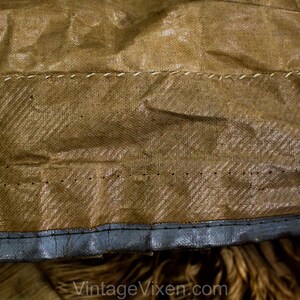 May include: Close-up of a section of vintage brown fabric with a textured, woven pattern. The fabric is stitched with white thread in a horizontal line. The bottom edge is trimmed with a dark grey material.