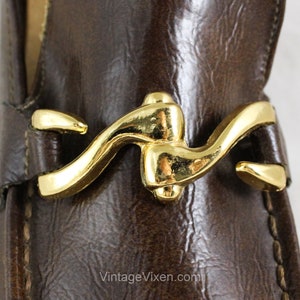 May include: Close-up of a brown leather loafer with a gold metal buckle. The buckle is a unique design with a curved shape.