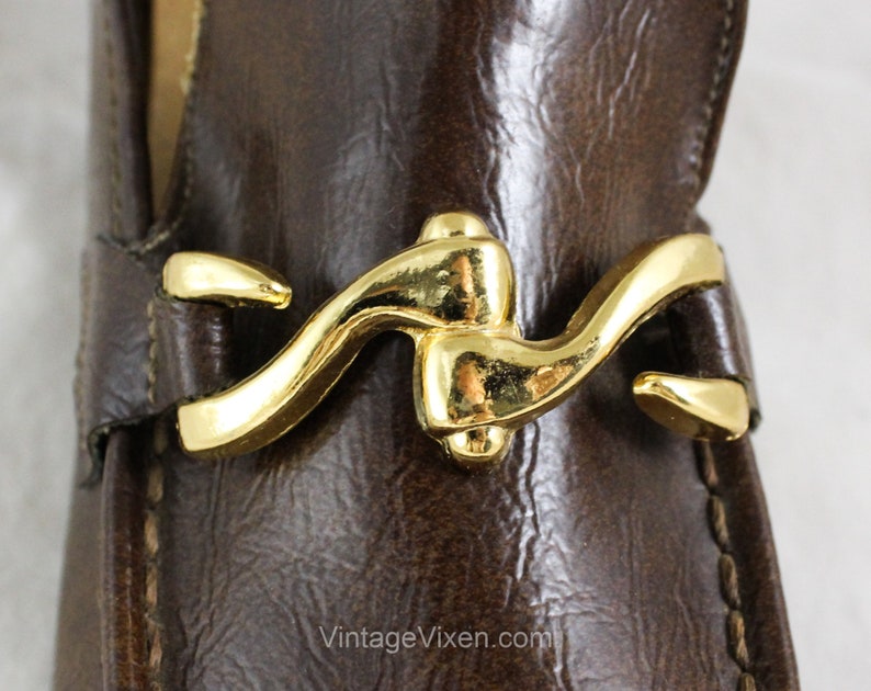 May include: Close-up of a brown leather loafer with a gold metal buckle. The buckle is a unique design with a curved shape.