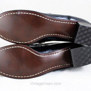 May include: A pair of black leather heeled shoes with a brown leather sole and a white stitch detail around the edge. The heels are block heels with a textured pattern.