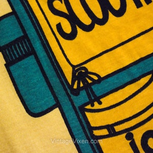 Puede incluir: Camiseta amarilla con un gr&aacute;fico negro y verde azulado de una mochila con el texto "School is"