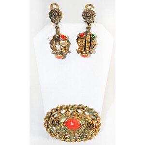 Puede incluir: Un conjunto de joyas doradas con piedras rojas y verdes. El conjunto incluye un par de pendientes colgantes y un broche.