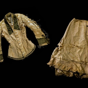 May include: A tan-coloured, distressed jacket and skirt set. The jacket has long sleeves with dark green accents and a notched collar. The skirt is long and flowing, with a ruffled hem. Both pieces show signs of wear.