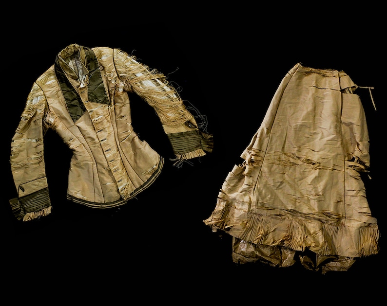 May include: A tan-coloured, distressed jacket and skirt set. The jacket has long sleeves with dark green accents and a notched collar. The skirt is long and flowing, with a ruffled hem. Both pieces show signs of wear.