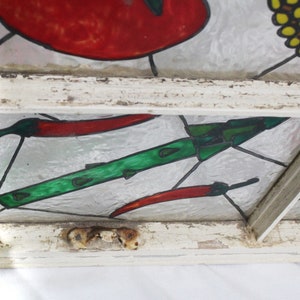 May include: A stained glass window pane with a white painted wooden frame. The glass features a green and black rocket, red chilli peppers, and a yellow and black flower.