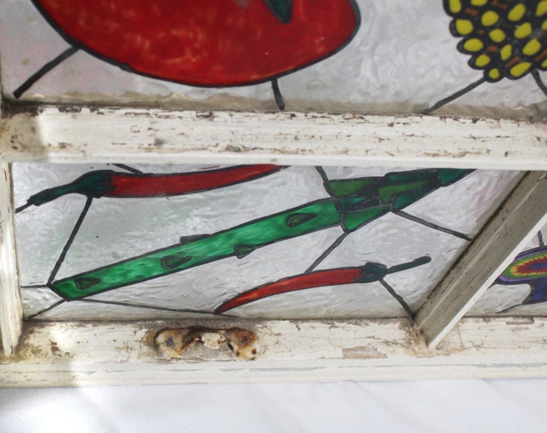 May include: A stained glass window pane with a white painted wooden frame. The glass features a green and black rocket, red chilli peppers, and a yellow and black flower.