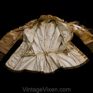 May include: A vintage cream-coloured jacket with a fitted waist and long sleeves. The jacket features a decorative belt, fringe detailing, and dark trim. The interior lining is a lighter cream colour. The jacket is displayed against a black background.