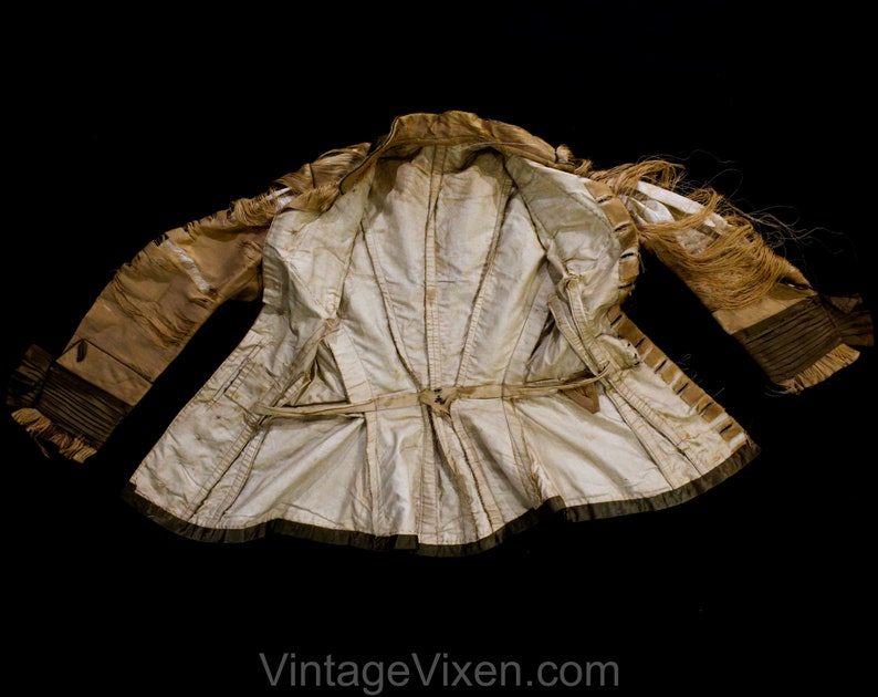 May include: A vintage cream-coloured jacket with a fitted waist and long sleeves. The jacket features a decorative belt, fringe detailing, and dark trim. The interior lining is a lighter cream colour. The jacket is displayed against a black background.