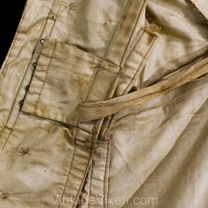 May include: Close-up of a vintage, off-white garment with visible wear and staining. Features include a pocket, reinforced seams, and a tie. The fabric shows signs of age and use, with dark stitching and metal eyelets.