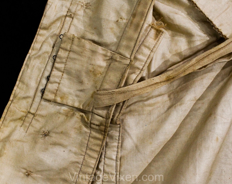 May include: Close-up of a vintage, off-white garment with visible wear and staining. Features include a pocket, reinforced seams, and a tie. The fabric shows signs of age and use, with dark stitching and metal eyelets.