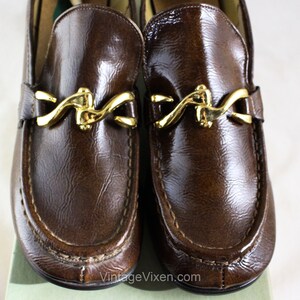 May include: A pair of brown leather loafers with gold metal buckles. The loafers have a pointed toe and a stitched seam around the edge.