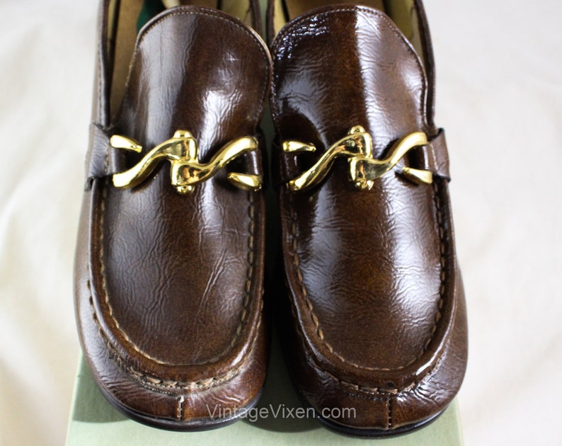 May include: A pair of brown leather loafers with gold metal buckles. The loafers have a pointed toe and a stitched seam around the edge.