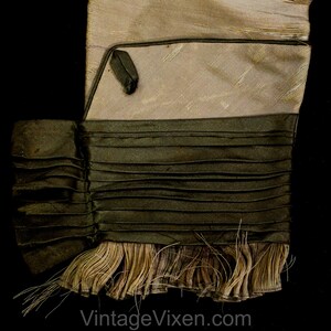 May include: A close-up of a vintage textile with a layered design. The top section is a light beige colour, followed by a dark olive green pleated section. The bottom features a fringe of beige threads against a black background.