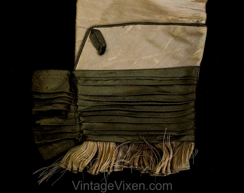 May include: A close-up of a vintage textile with a layered design. The top section is a light beige colour, followed by a dark olive green pleated section. The bottom features a fringe of beige threads against a black background.