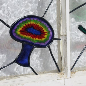 May include: A stained glass window panel featuring a colourful mushroom with a blue stem and a rainbow-coloured cap. The mushroom is set against a white background.
