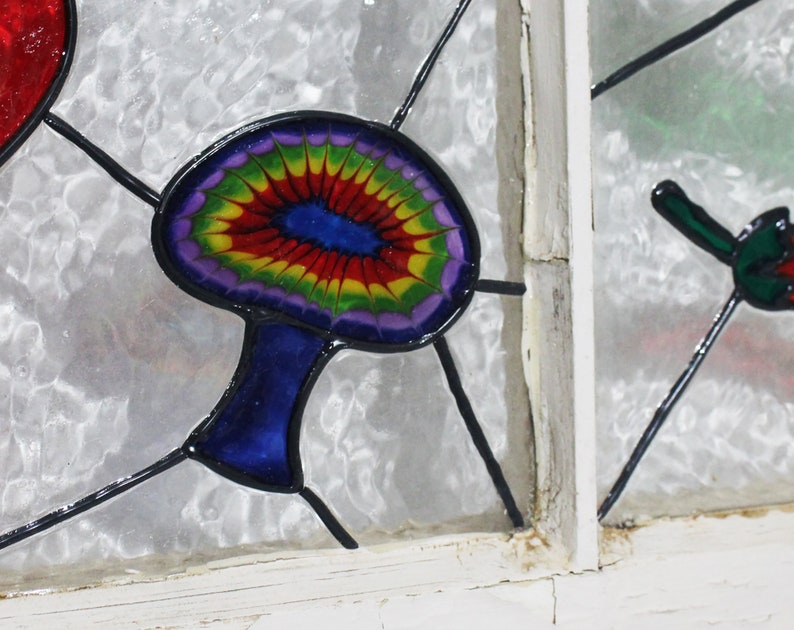 May include: A stained glass window panel featuring a colourful mushroom with a blue stem and a rainbow-coloured cap. The mushroom is set against a white background.
