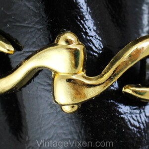 May include: Close-up of a gold-toned metal buckle with a curved design. The buckle is attached to a black leather strap.