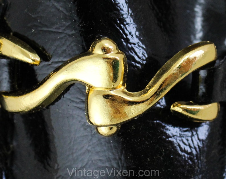 May include: Close-up of a gold-toned metal buckle with a curved design. The buckle is attached to a black leather strap.