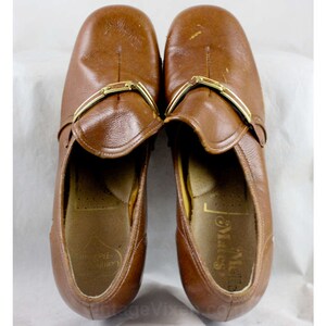 May include: A pair of brown leather slip-on shoes with gold buckles. The shoes have a slightly pointed toe and a low heel. The brand name "Miss" is visible on the inside of the right shoe.