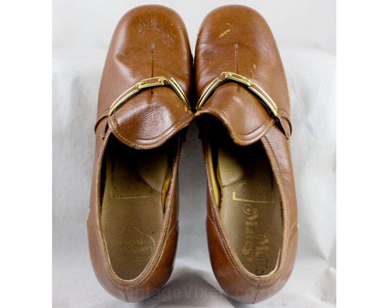 May include: A pair of brown leather slip-on shoes with gold buckles. The shoes have a slightly pointed toe and a low heel. The brand name "Miss" is visible on the inside of the right shoe.
