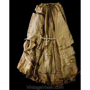 May include: A vintage, tan dress with a distressed, textured look. The dress has a layered design with frayed edges and a gathered waist, secured by a white band. The hemline is fringed. The text "VintageVixen.com" is visible. The dress appears to be made of a light fabric.