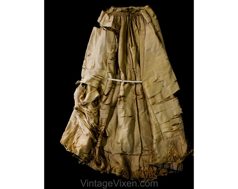 May include: A vintage, tan dress with a distressed, textured look. The dress has a layered design with frayed edges and a gathered waist, secured by a white band. The hemline is fringed. The text "VintageVixen.com" is visible. The dress appears to be made of a light fabric.