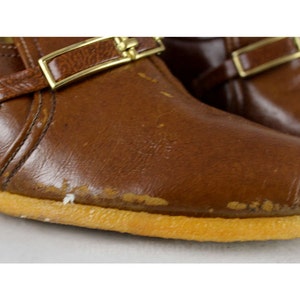 May include: A pair of brown leather shoes with gold buckles. The shoes have a rubber sole and a slightly pointed toe.