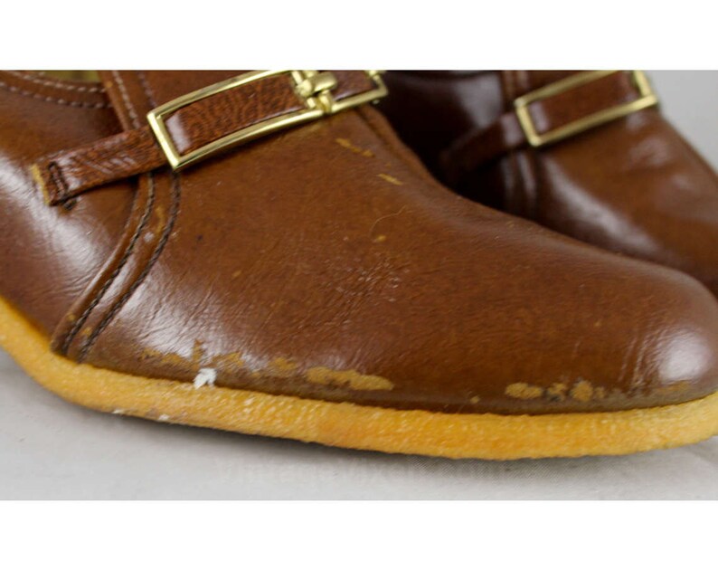 May include: A pair of brown leather shoes with gold buckles. The shoes have a rubber sole and a slightly pointed toe.