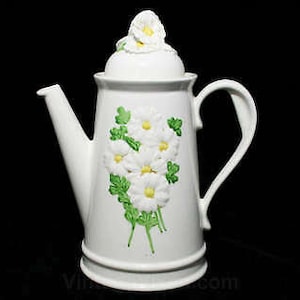 May include: White ceramic teapot with a floral design. The teapot has a lid with a single white flower on top. The floral design features white flowers with green leaves.