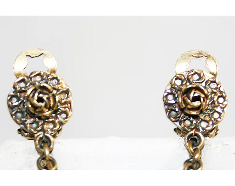 Puede incluir: Un par de pendientes de color dorado con un dise&ntilde;o de rosa y una cadena. Los pendientes tienen un peque&ntilde;o c&iacute;rculo abierto en la parte superior y un c&iacute;rculo m&aacute;s grande con una rosa en el centro. La rosa est&aacute; rodeada de peque&ntilde;as piedras transparentes.