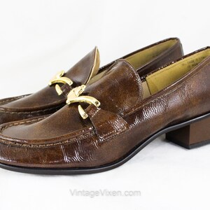 May include: Brown leather heeled loafers with a gold buckle detail. The loafers have a pointed toe and a low block heel.