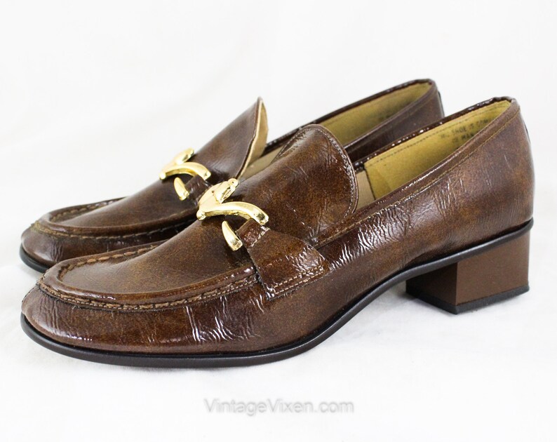 May include: Brown leather heeled loafers with a gold buckle detail. The loafers have a pointed toe and a low block heel.