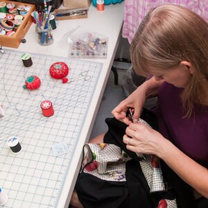 May include: A woman sits at a table sewing a black and white fabric with a floral pattern. She is using a needle and thread and has a variety of sewing supplies on the table, including spools of thread, thimbles, and scissors.