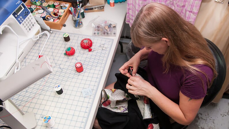 May include: A woman sits at a table sewing a black and white fabric with a floral pattern. She is using a needle and thread and has a variety of sewing supplies on the table, including spools of thread, thimbles, and scissors.