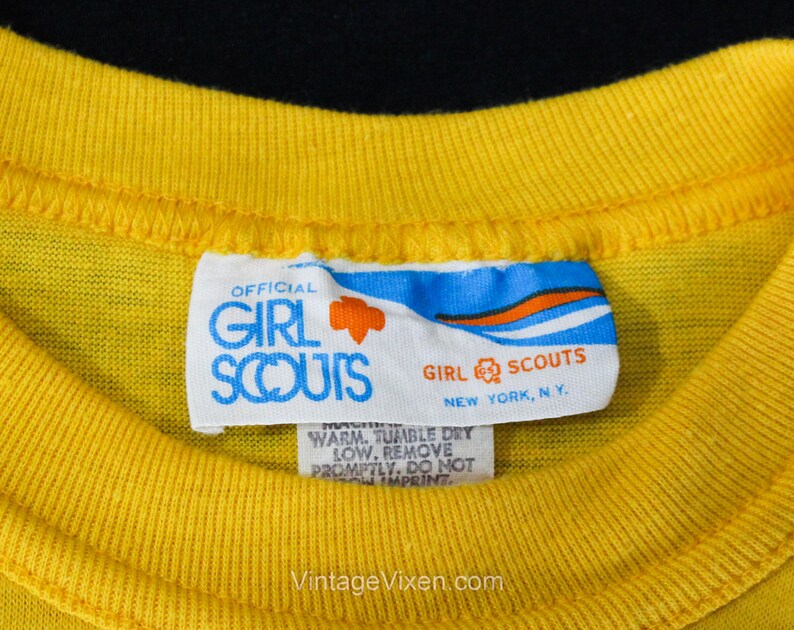 Puede incluir: Una etiqueta blanca con detalles azules y naranjas que dice "OFFICIAL GIRL SCOUTS NEW YORK, NY."