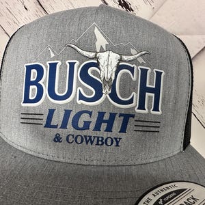 May include: Gray trucker hat with a black mesh back. The front features the words "BUSCH LIGHT & COWBOY" in blue and white, with a bull skull graphic and mountain range.