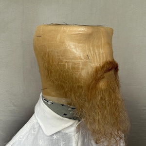 May include: A mannequin head covered in tan fabric, with a long, reddish-brown beard. The mannequin wears a white shirt with a floral pattern and a patterned neckerchief.