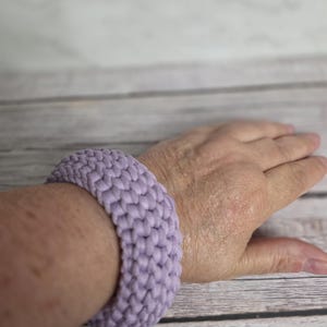 May include: A lavender-colored, woven bracelet is worn on a person's wrist. The bracelet is thick and appears to be made of a soft, textured material. The background is a light-colored wooden surface.