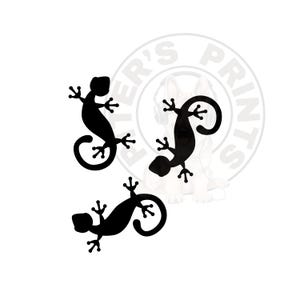 May include: Three black gecko silhouettes with curled tails and detailed feet are arranged on a white background. The geckos are in various poses, creating a dynamic and artistic design. The image has a clean, minimalist aesthetic.