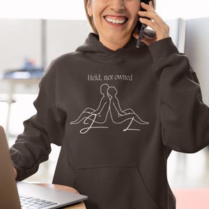 Puede incluir: Sudadera con capucha de color chocolate oscuro con cordones. La sudadera presenta el texto "Held, not owned" y un dibujo lineal de dos figuras. La imagen también incluye un portátil y un sello de "Quality Guarantee".