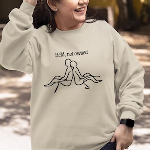 Puede incluir: Sudadera de color arena con el texto "Held, not owned" y un dibujo lineal negro de dos figuras. La sudadera tiene mangas largas y cuello redondo.