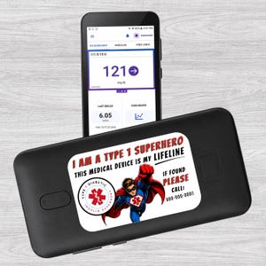 Può includere: Un dispositivo medico nero con un adesivo bianco che recita "I AM A TYPE 1 SUPERHERO THIS MEDICAL DEVICE IS MY LIFELINE". Uno smartphone mostra una lettura della glicemia di 121 mg/dL.