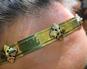Könnte beinhalten: Ein goldfarbenes Metall-Stirnband mit einer gehämmerten Textur auf dem Band und gleichmäßig verteilten, erhabenen quadratischen Ornamenten mit detaillierten blattartigen Verzierungen. Das Band ist dünn und flexibel für den Stirnbereich.