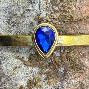 May include: A gold-coloured metal barrette featuring a teardrop-shaped, faceted, deep blue gemstone. The gemstone is set in a gold bezel. The barrette is set against a textured, mossy background.