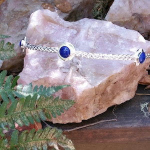 May include: A silver-toned metal headband with a woven design, featuring three circular settings with vibrant blue gemstones. The headband is displayed on a light pink rock, with green ferns and fallen leaves in the background.