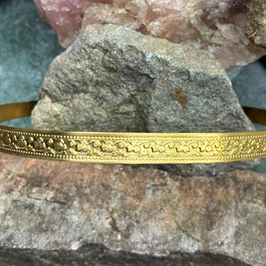 May include: A gold-colored bangle bracelet with an intricate floral pattern. The bracelet is displayed on a natural stone surface, showcasing its detailed design and metallic sheen. The bangle's circular shape and decorative elements are clearly visible.