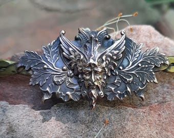 May include: A detailed silver-toned metal Green Man pendant with a face, wings, and leaf details. The pendant is attached to a braided green and gold band. The piece is set against a natural, textured background.