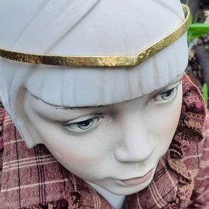 Könnte beinhalten: Ein weißer Porzellan-Puppenkopf mit einem goldenen Stirnband. Die Puppe trägt ein rot-braunes kariertes Kleid mit einem Rüschenkragen.